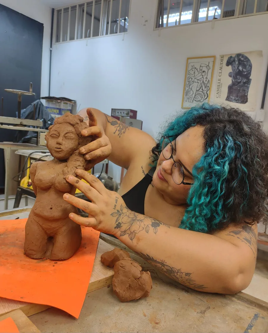 Lucy modelando com as mãos uma escultura de argila de figura feminina, na bancada do ateliê