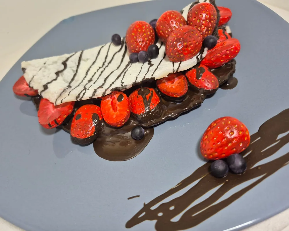 Close da escultura de crepe com morangos pintados um a um, mirtilos e calda de chocolate, num prato azul