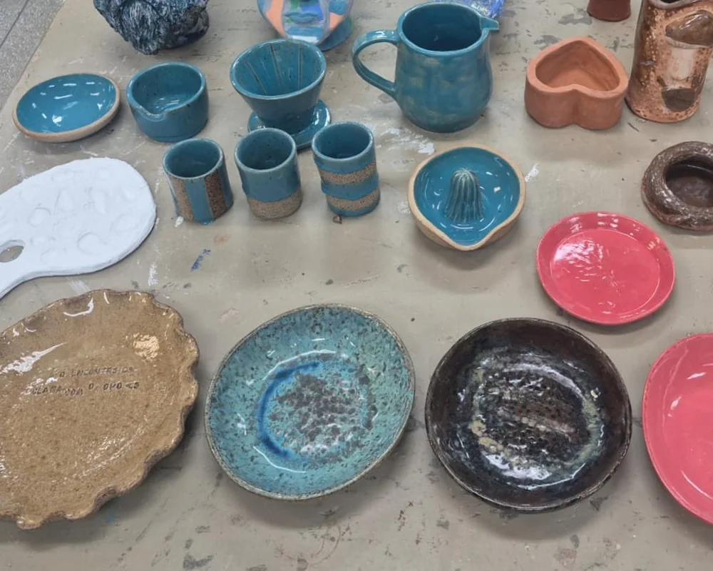 Conjunto de peças de cerâmica esmaltadas reunidas na bancada do ateliê: canecas, jarra, coador, espremedor, tigelas e pratos