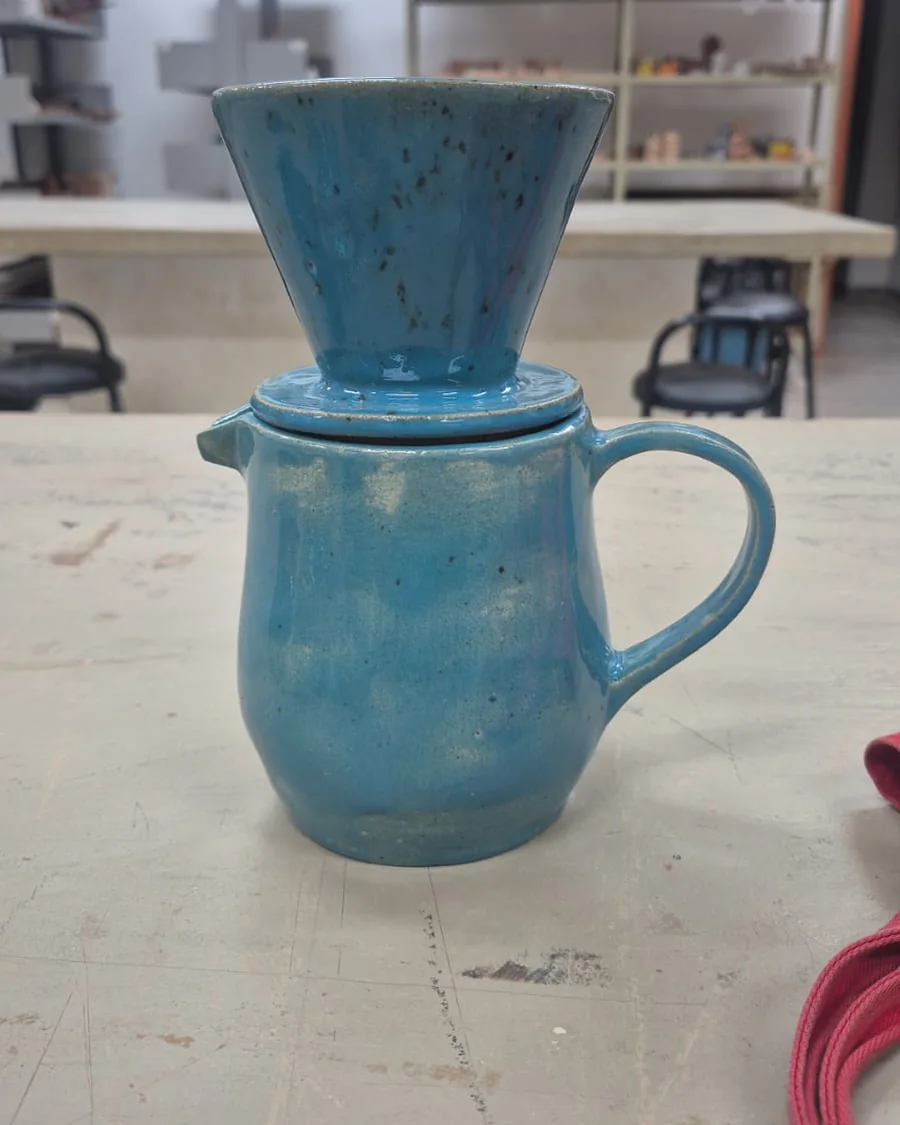 Conjunto de coador e jarra de cerâmica para café, com esmalte azul salpicado, sobre a bancada do ateliê
