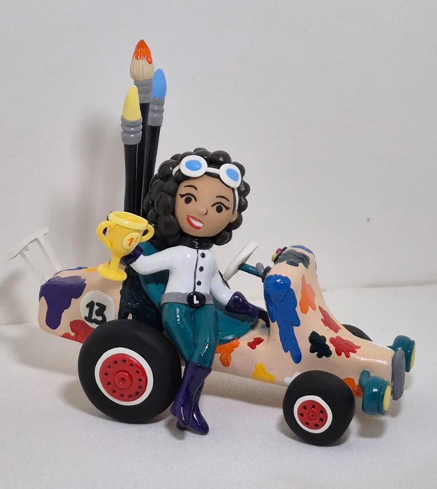 Escultura autoral em biscuit de uma menina piloto sentada em um kart pintado com respingos coloridos, segurando um troféu, com pincéis gigantes atrás
