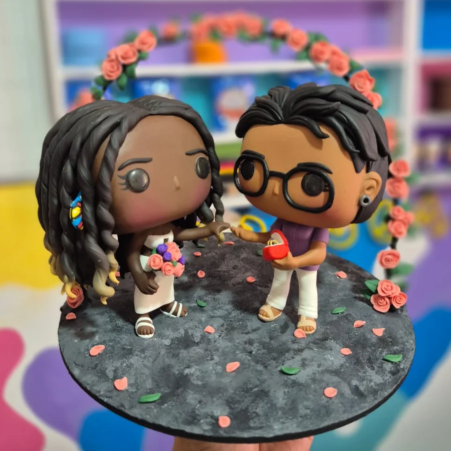 Pop de casal: noiva com buquê e noivo de óculos sobre base com pétalas e arco de flores, feito sob encomenda