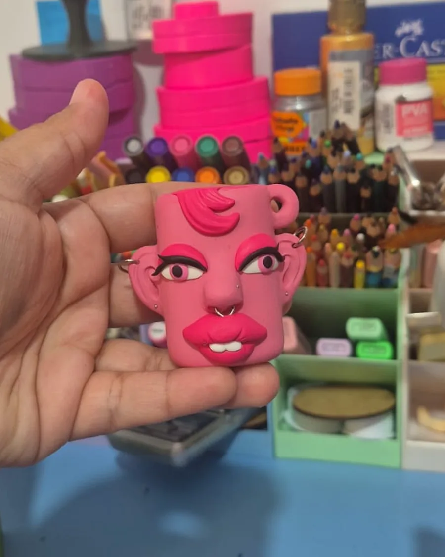Case de isqueiro em biscuit no formato de um rosto rosa, com piercings na orelha e no nariz e batom marcado, segurado na mão sobre a bancada do ateliê