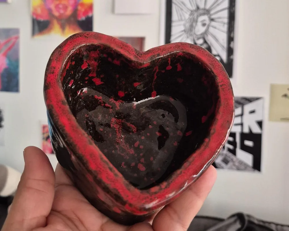 Bowl de cerâmica em formato de coração, com esmalte vermelho salpicado sobre fundo preto, segurado na palma da mão