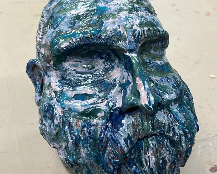 Escultura autoral de uma cabeça barbada, pintada à mão em azul, verde e branco, sobre a bancada do ateliê